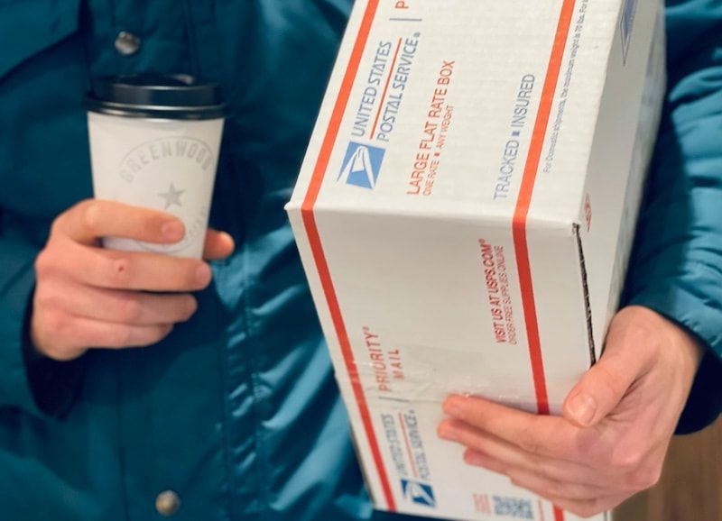 Person holding a coffee cup and a USPS Priority Mail box in a blue jacket