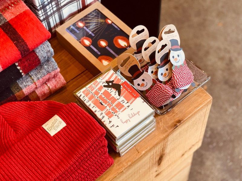 Folded sweaters and knitted items displayed on a wooden table with magazines and red plaid fabric.