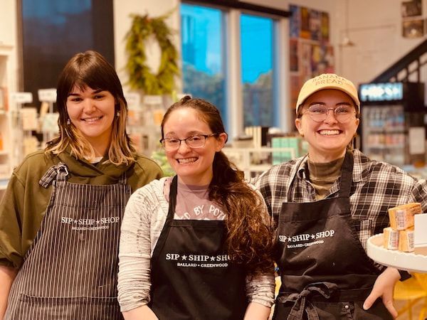 Three smiling bakery staff in aprons stand behind the counter holding a tray of pastries.