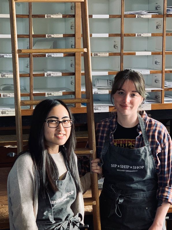 Two people smiling in front of a wall of wooden cubby shelves in a café or library.