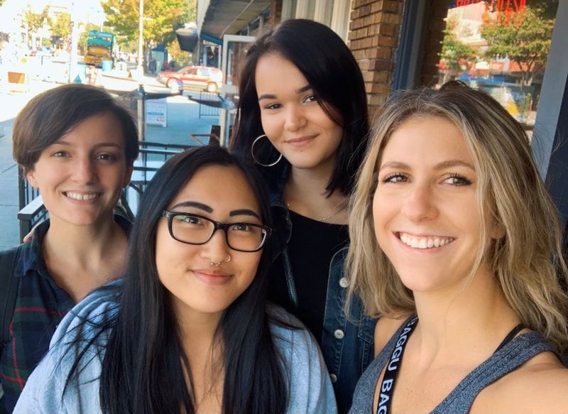 Four smiling people posing outdoors on a sunny street, with storefronts and parked cars behind them.