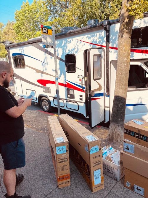 Man loading boxes beside a white RV with red and blue stripes in a tree-lined parking area