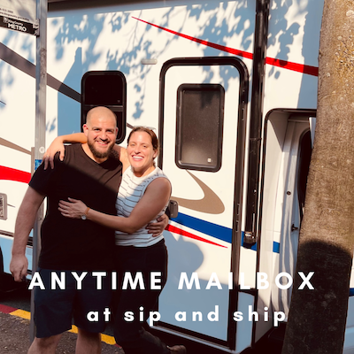 Two smiling people standing beside an Anytime Mailbox trailer with “at sip and ship” text.