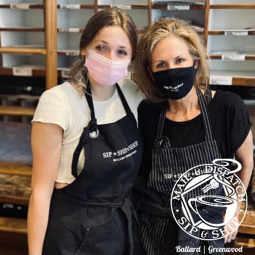 Two bakery staff in aprons and masks smiling behind a counter