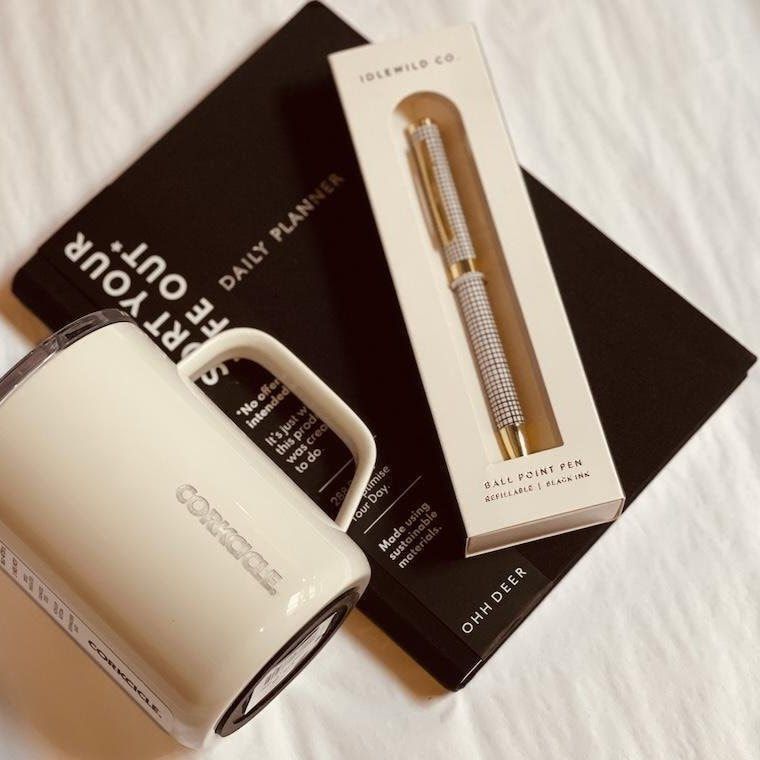 White mug, black notebook, and gold pen on a soft white surface.