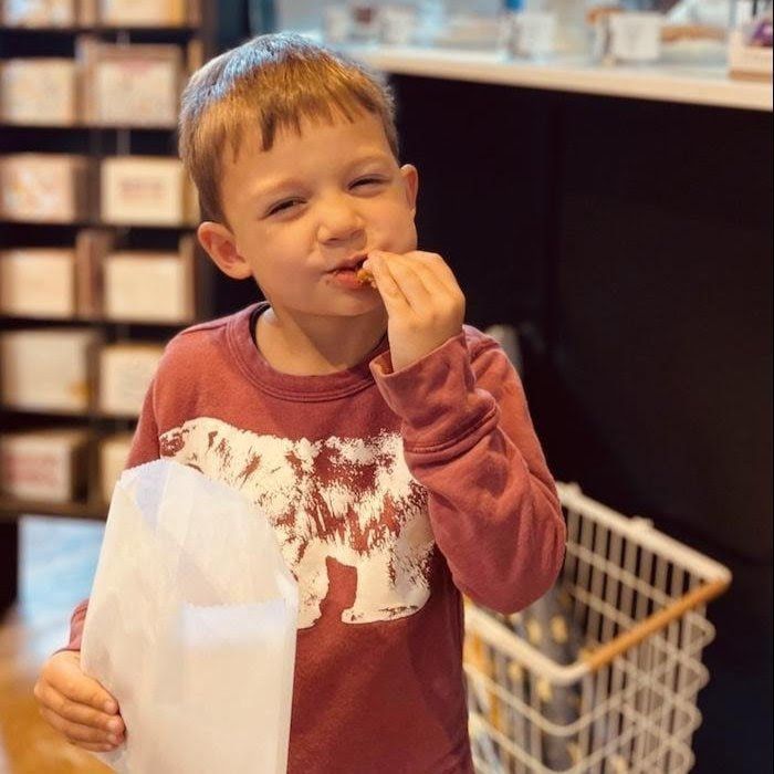 Child eating a snack in a store aisle, holding a white bag and wearing a maroon sweater