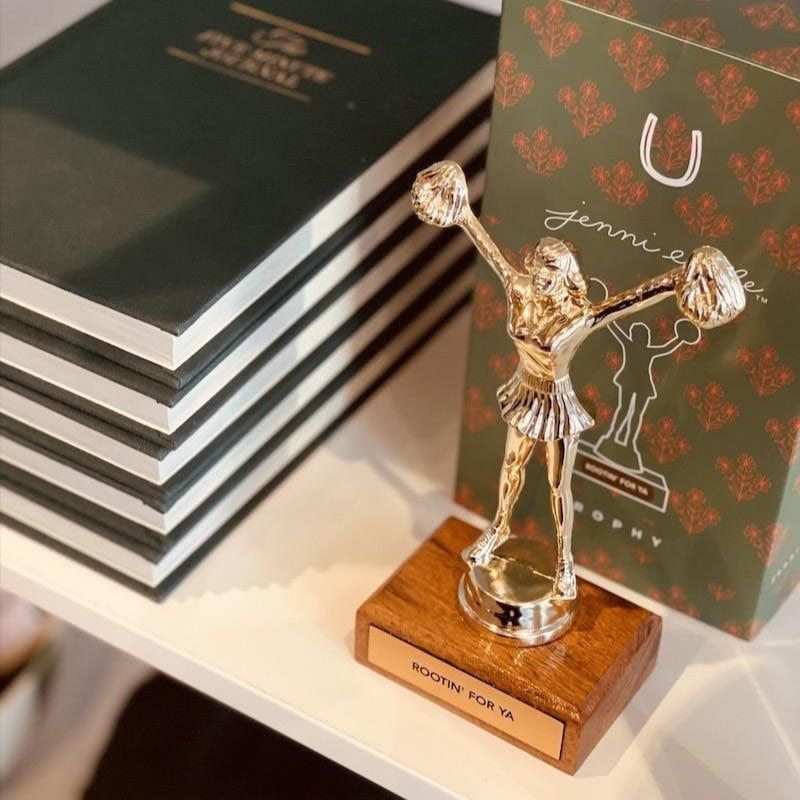 Gold cheer trophy on a wooden base beside stacked black books and a patterned award card on a shelf