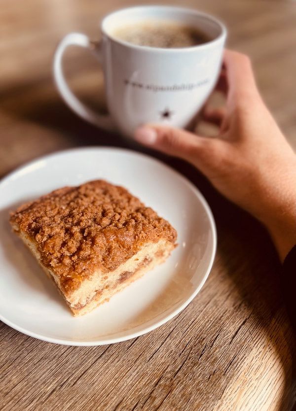 Hand holding a white mug beside a plate with a crumb-topped slice of cake on a wooden table