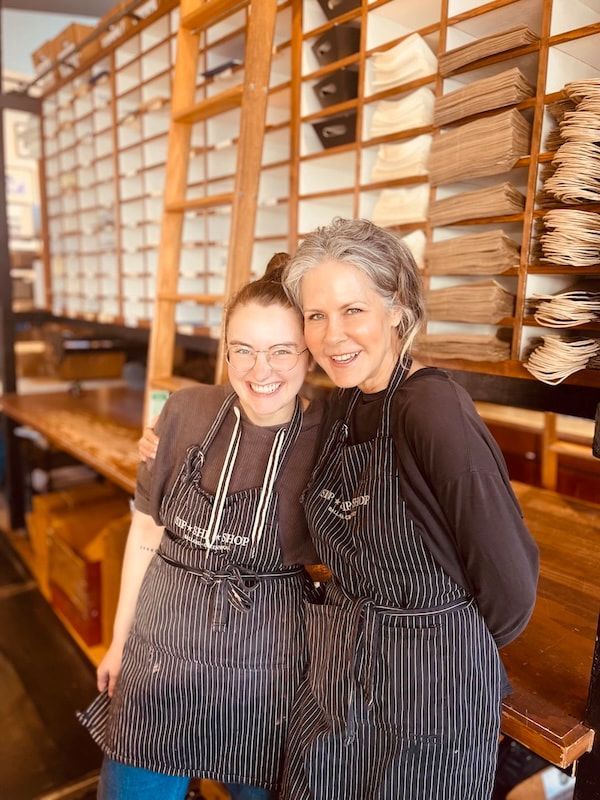 Two smiling people in striped aprons lean together in a bakery with shelves of stacked trays.
