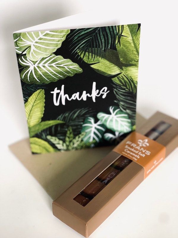 Thank-you card with tropical leaves beside a brown box of chocolates on a white surface