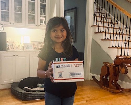 Young child holding a USPS package in a home living room with stairs in the background