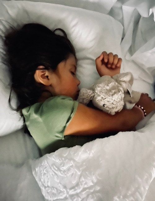 Child sleeping in white bedding, holding a small stuffed toy.