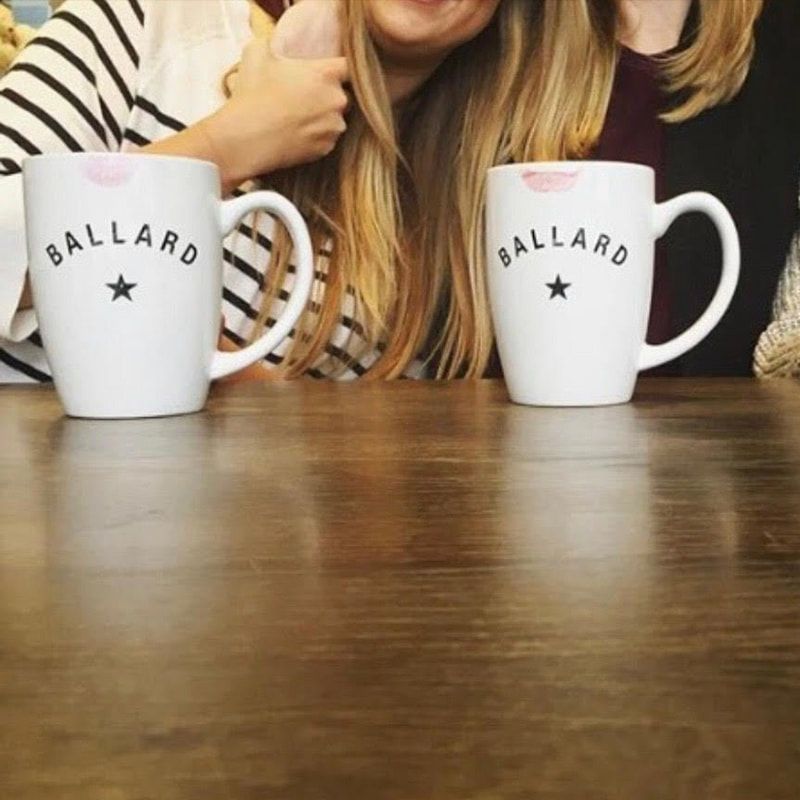 Two white Ballard mugs on a wooden table, with a person sitting behind them.