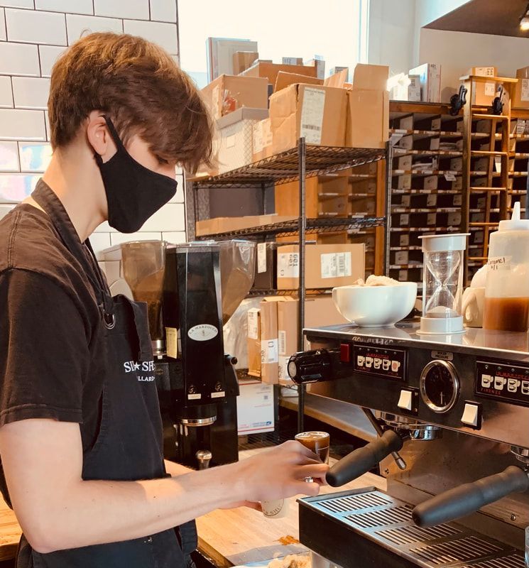 Barista in black apron and mask preparing espresso at a café counter with stacked boxes behind him