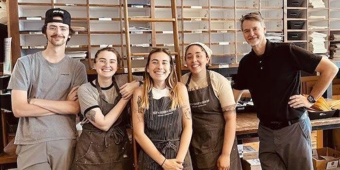 Five people posing in a bakery, smiling in front of shelves of baked goods.
