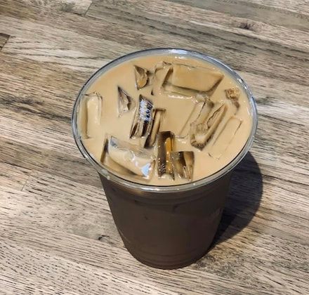 Glass of iced coffee on a wooden table, topped with ice cubes and light tan milk foam.