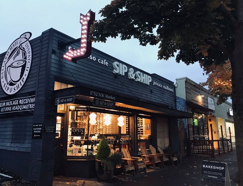 Corner café storefront at dusk with warm lights, black siding, and a glowing red arrow sign reading “SIP & SIP.”