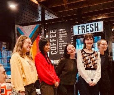 Five smiling people standing indoors in front of a coffee shop sign and a “FRESH” display.