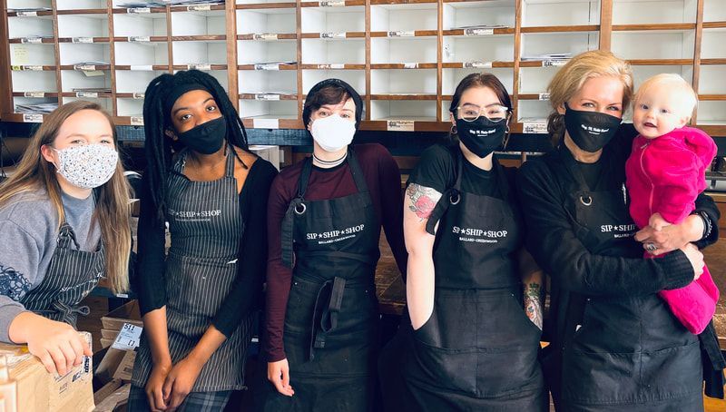 Five masked people in black aprons pose indoors; one holds a baby in pink.