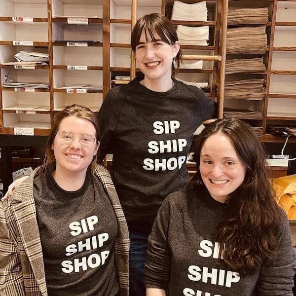 Three smiling people in matching “SIP SHIP SHOP” shirts standing in a shop with shelves of paper products.