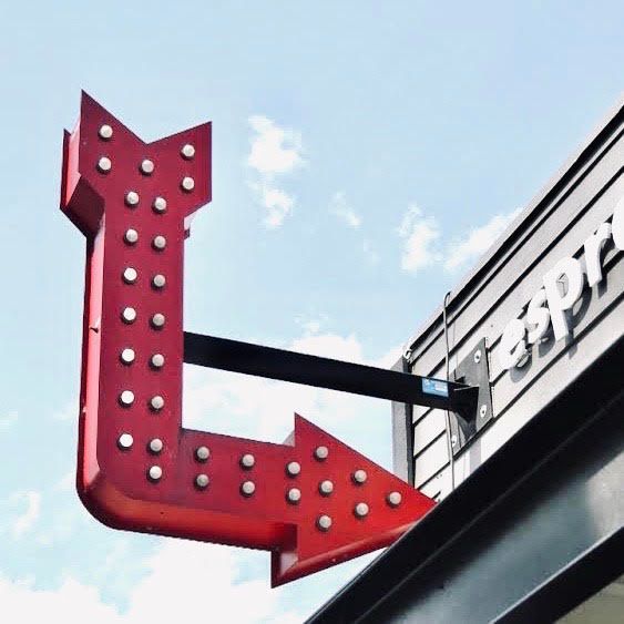 Red arrow-shaped hanging sign with white bulbs on a building exterior against a blue sky