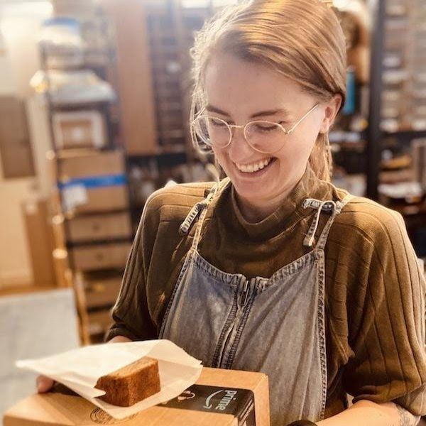 Smiling person in glasses and apron holding a box with a baked loaf in a warm bakery