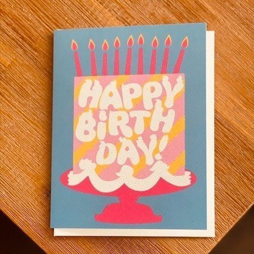 Birthday card with pink cake and candles on a blue background, reading “Happy Birthday.”