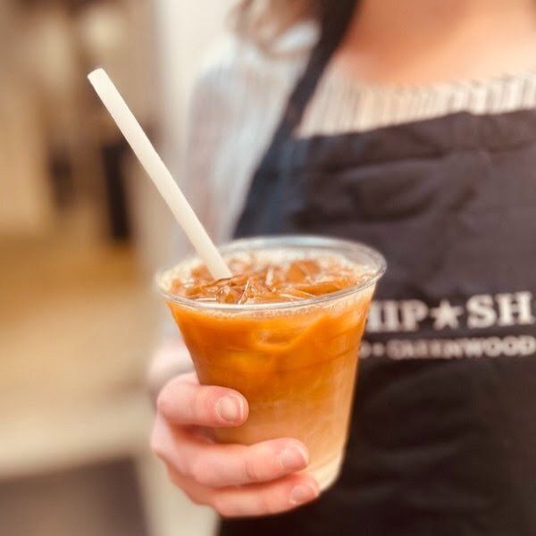 Person holding an iced orange drink in a clear plastic cup with a straw, wearing a dark apron