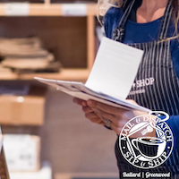 Shop worker in blue apron holding a clipboard with paper at a deli counter.