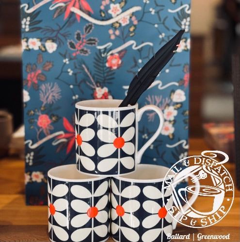 Black-and-white floral mug and saucer with an orange accent, holding a black spoon, in front of a blue floral backdrop