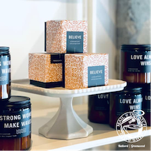 Gift boxes on a white pedestal, surrounded by dark mugs and tins with “love” and “believe” text