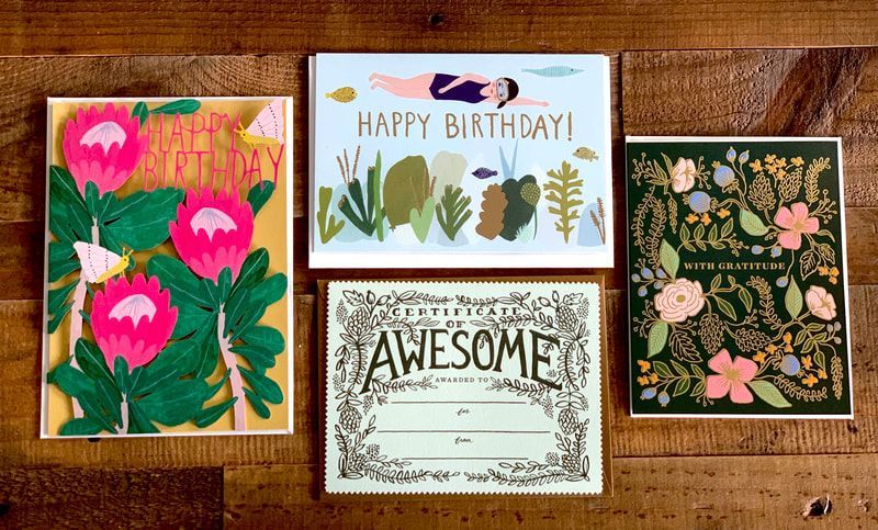 Four floral greeting cards, including “Happy Birthday!” and “Awesome,” arranged on a wooden surface.