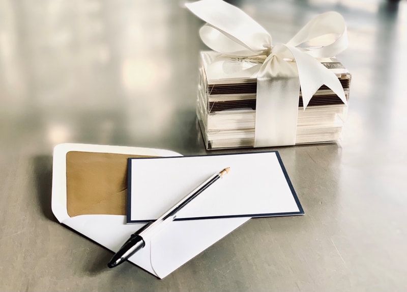 White gift boxes tied with ribbon beside a stack of envelopes and a pen on a silver surface