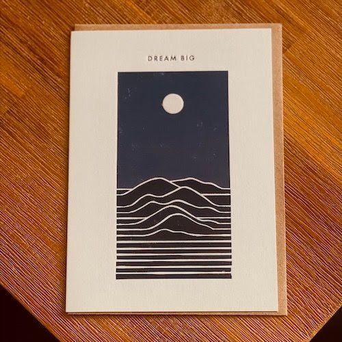 Minimalist poster with a moon over layered mountains, labeled “DREAM BIG,” on a wooden surface