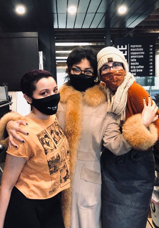 Three masked people posing together indoors, wearing winter coats and fur-trimmed hoods.