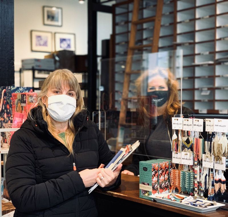 Masked shop clerk holding a tablet behind a jewelry display in a boutique