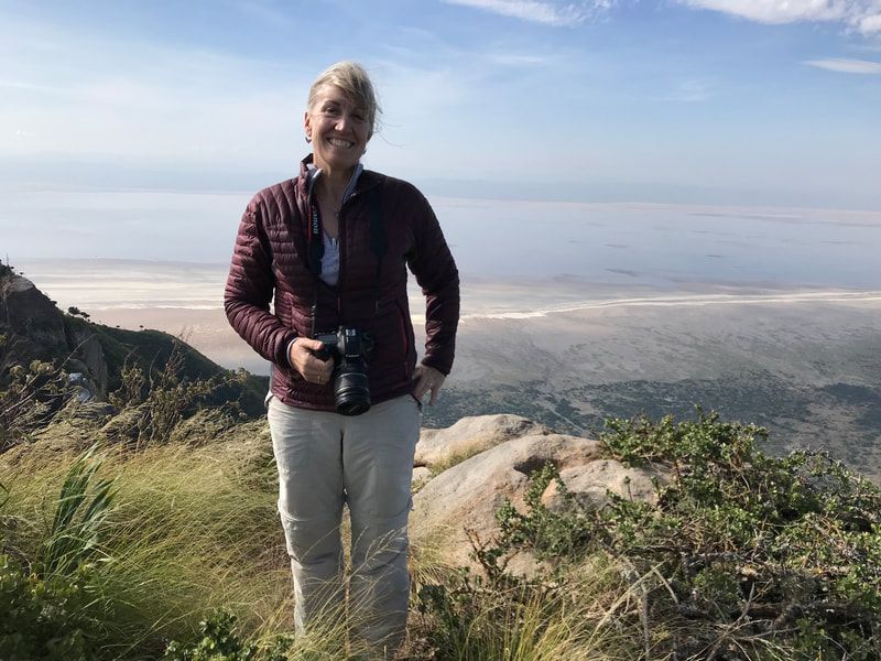 Person standing on a grassy cliff overlooking a beach, holding a camera under a sunny sky