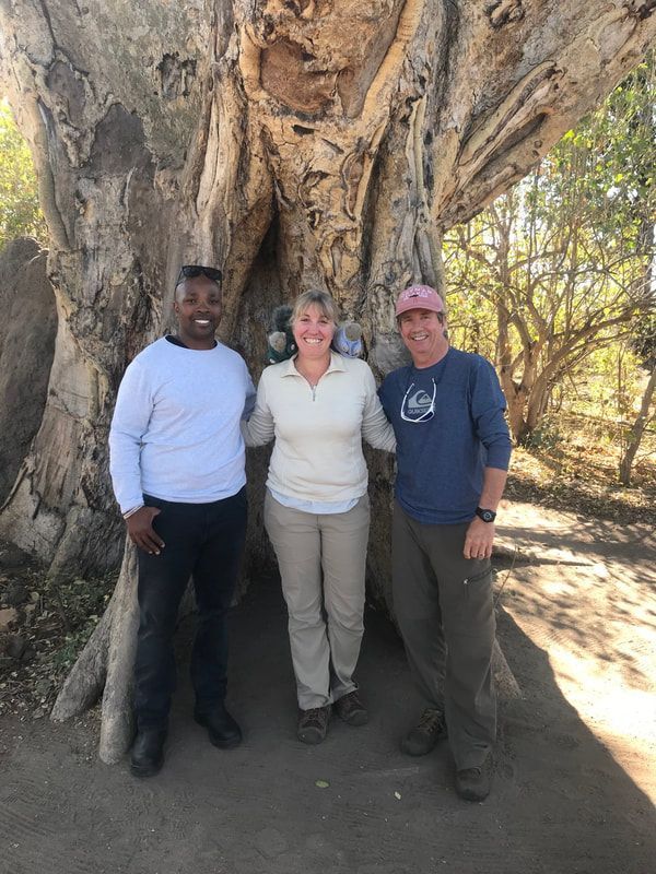 Three people standing together under a large tree in a wooded area, smiling at the camera.