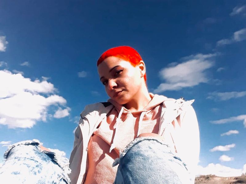 Person in red cap and white jacket looking down against a blue sky with clouds, wearing denim jeans