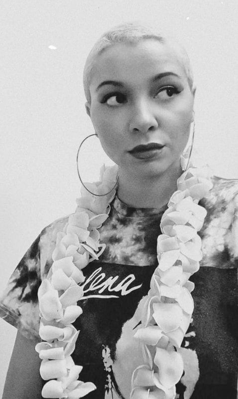 Person in a black top and flower lei, wearing hoop earrings, looking to the side against a light background.