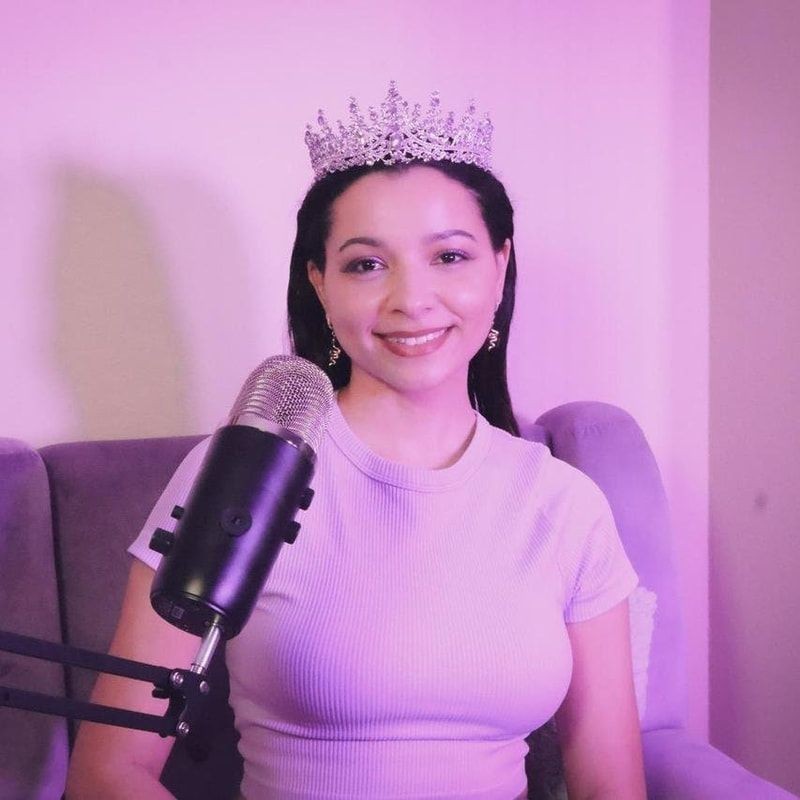 Smiling woman wearing a tiara beside a microphone in a pink-lit room