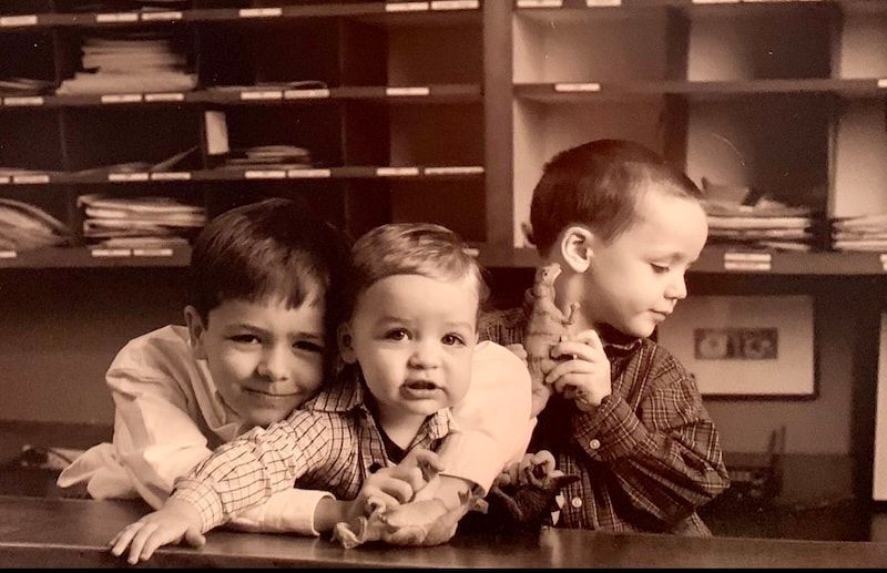 Three children lean on a table, smiling and looking in different directions in a classroom.