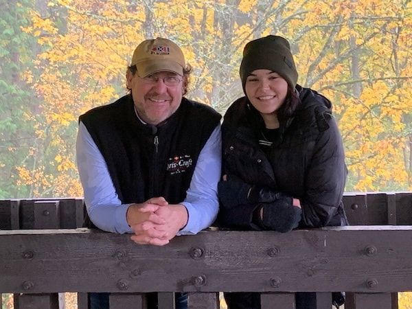Two people in jackets and hats lean on a wooden railing with autumn trees behind them.