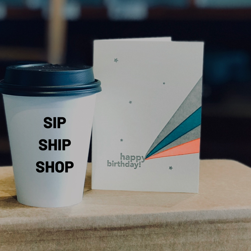 White birthday card beside white coffee cup on wooden table, with “SIP SHIP SHOP” text and colored geometric accents.