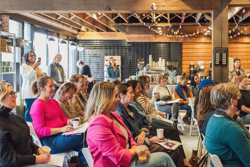 Audience listening in a bright conference room, seated on folding chairs with drinks and laptops