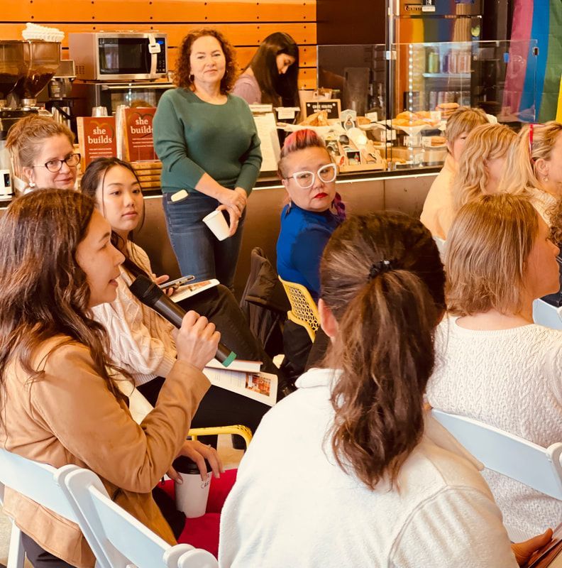 Audience seated in a café, one woman speaking into a microphone during a small discussion event