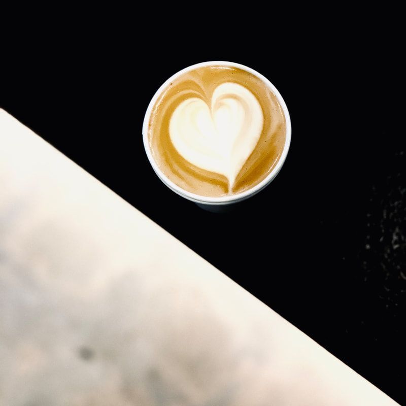 Cup of latte with heart-shaped foam art on a black table, beside a blurred white surface.