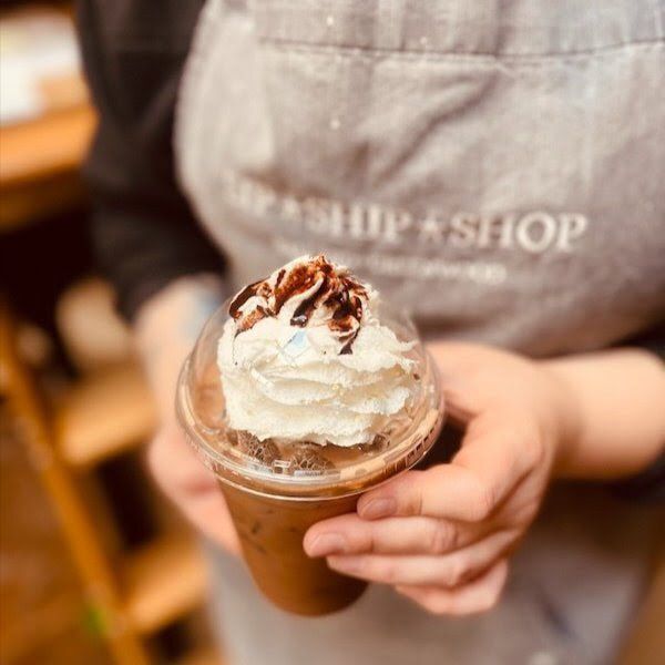 Person holding iced coffee topped with whipped cream and chocolate drizzle in a café apron