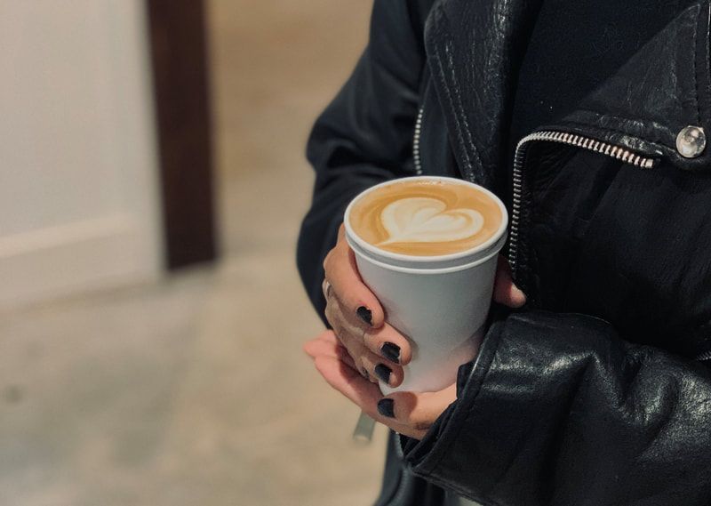 Hand holding a small gray cup of latte with heart-shaped foam art, wearing a black jacket.