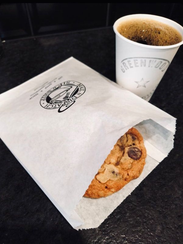 Cookie in white paper bag beside a paper cup of coffee on a dark counter.